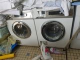 une machine à laver et un sèche-linge dans une pièce où il y a beaucoup de vêtements par terre