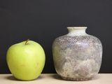 a green apple next to a gray vase on a table