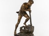 une statue en bronze représentant un homme tenant une lance, assis sur un rocher