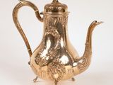 une théière ancienne en métal argenté, ornée de motifs élaborés, posée sur un socle
