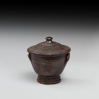 un vase marron muni d'un couvercle et d'une poignée, posé sur une surface blanche