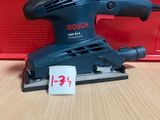 une ponceuse orbitale Bosch posée sur une table, à côté d'une boîte rouge