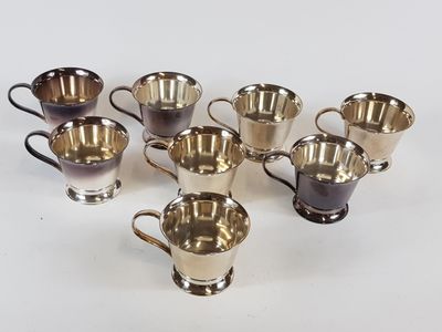 six tasses en métal argenté et une tasse en métal argenté posées sur un fond blanc