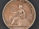 une image représentant une médaille d'argent sur laquelle figure une femme assise sur une boîte