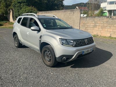 La Dacia blanche est garée sur un parking, près d'un mur de briques.