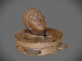 sculpture représentant une tête posée sur un socle en bois, sur fond gris