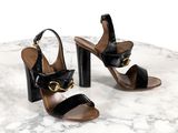 des sandales en cuir noir ornées de boucles dorées, posées sur une table en marbre