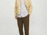 un homme debout devant un fond blanc, vêtu d'une veste beige et d'un pantalon marron
