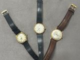 trois montres différentes posées sur un fond gris, dont une dorée et une noire