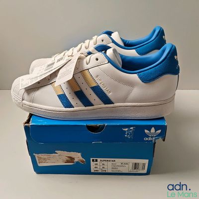 des chaussures Adidas Superstar blanches et bleues posées sur une boîte bleue