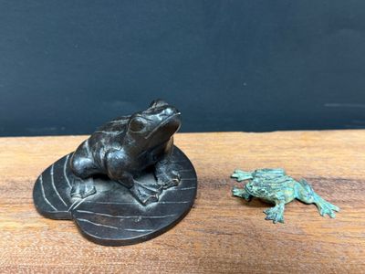 a small bronze frog sitting on top of a leaf next to a small frog