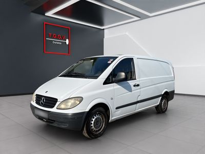 La camionnette blanche est garée dans une grande pièce où se trouve une affiche rouge accrochée au mur.