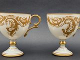 two white and gold cups with gold designs on them on a gray surface