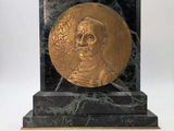 une image représentant un médaillon en or orné du visage d'un homme, posé sur un socle en marbre