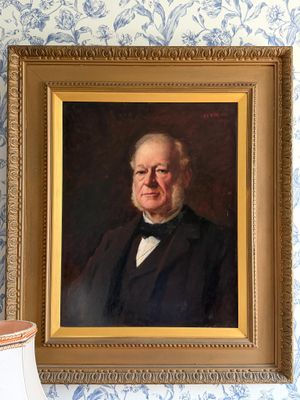 portrait d'un homme vêtu d'un manteau noir et d'un nœud papillon, devant un mur recouvert d'un papier peint bleu et blanc
