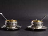 two silver cups and saucers with spoons sitting on a black surface