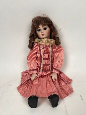 une poupée ancienne assise sur une surface blanche, vêtue d'une robe rose et de chaussures noires