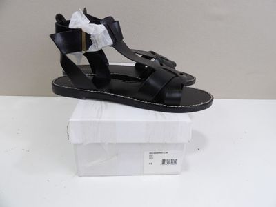 Des sandales en cuir noir ornées de nœuds sur les orteils sont présentées dans une boîte blanche.