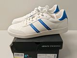 Baskets adidas Originals blanches et bleues, ornées de bandes bleues, posées sur une boîte
