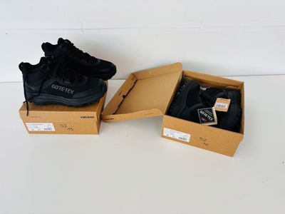 Deux paires de baskets noires se trouvent dans une boîte posée sur une surface blanche