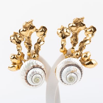 une paire de boucles d'oreilles en forme de coquillage, dorées et blanches, posées sur un socle blanc