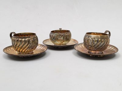 trois tasses et soucoupes en métal argenté posées sur des soucoupes, sur une table