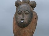 sculpture représentant un visage posée sur un socle en bois, avec un ciel gris en arrière-plan
