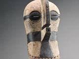une image représentant un masque en bois posé sur un socle, sur fond gris