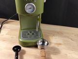 a green coffee maker on a wooden table with a measuring spoon