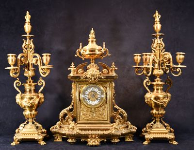 une horloge en or posée sur une table, à côté de trois chandeliers