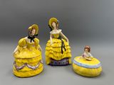 trois figurines en porcelaine représentant des femmes vêtues de robes jaunes et coiffées de chapeaux