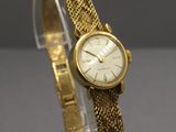 une photo d'une montre en or avec un bracelet maillé, posée sur un socle transparent