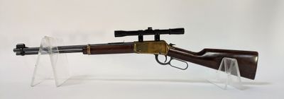 un vieux fusil avec un canon en bois et une lunette de visée, posé sur un socle en acrylique transparent
