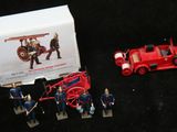 un camion de pompiers rouge en jouet et quelques figurines posées devant une boîte