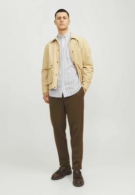 un homme debout devant un fond blanc, vêtu d'une veste beige et d'un pantalon marron