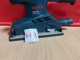 une ponceuse orbitale Bosch posée sur une table en bois, avec une boîte rouge en arrière-plan