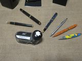 several pens, a brush, a pen holder, and a cell phone on a table