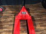 un chariot élévateur rouge posé sur un plancher en bois, à côté d'une paire de pieds