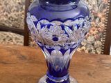 purple glass vase on wooden table in front of wallpapered wall