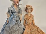 two dolls of women in dresses and hats are standing next to each other