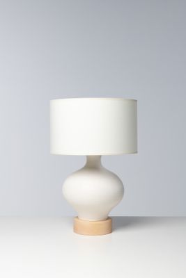 La lampe de table est blanche ; elle est dotée d'un pied en bois et d'un abat-jour blanc.