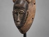 un masque en bois muni de cornes, posé sur un socle, sur fond gris