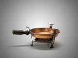 une vieille casserole en cuivre avec une poignée en bois, posée sur un support