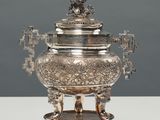 une théière ancienne en métal argenté, posée sur un support, sur une table blanche