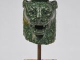 une photo représentant une sculpture verte représentant une tête, posée sur un socle en bois