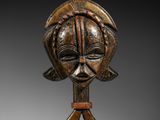 un masque en bois représentant un visage et deux mains, posé sur un socle