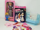 une poupée Barbie dans une boîte rose, à côté d'une boîte contenant des ciseaux et d'autres objets