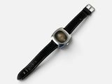 une photo d'une montre posée sur une surface blanche, avec un bracelet noir