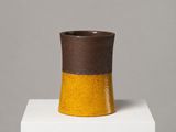 vase en céramique marron et jaune posé sur un socle blanc, sur fond gris