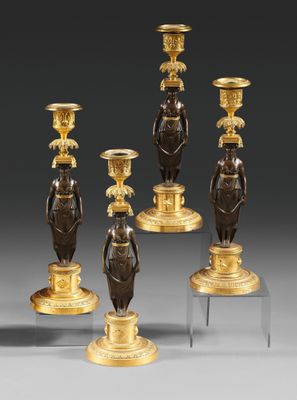 quatre bougeoirs en bronze ornés de figurines, posés sur des socles, sur un fond gris